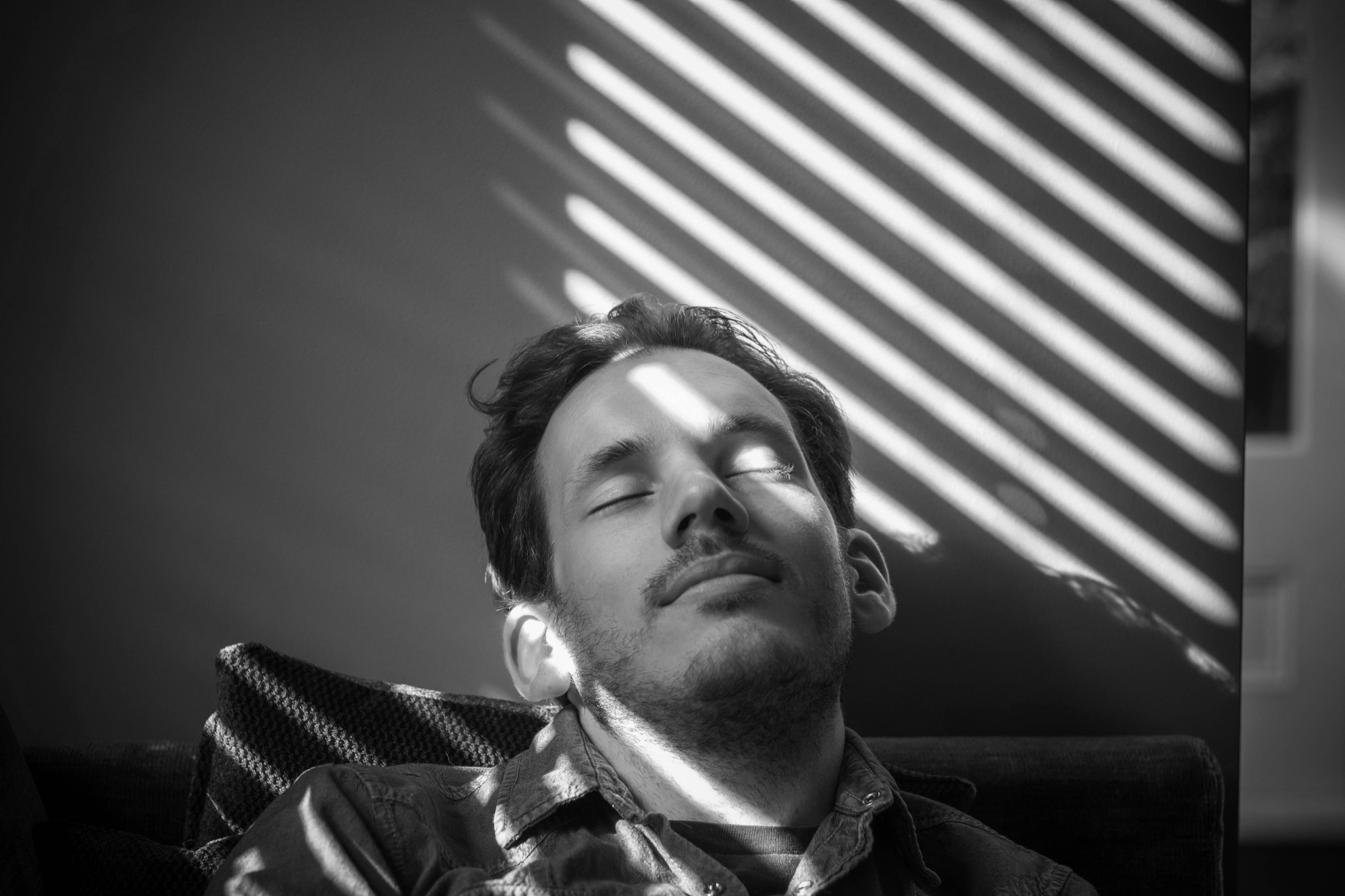 A man sitting back with his eyes closed, sunlight through the blinds across his face